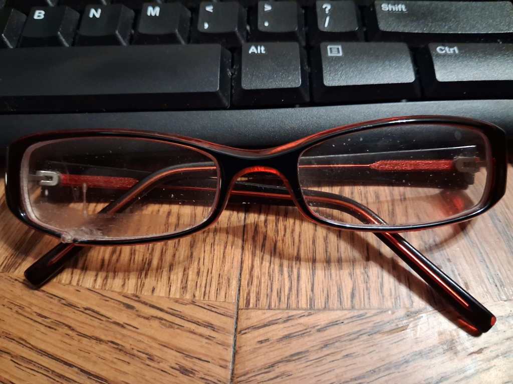 pair of glasses on a wooden table in front of a keyboard that has superglue on the bottom of the lens on the left